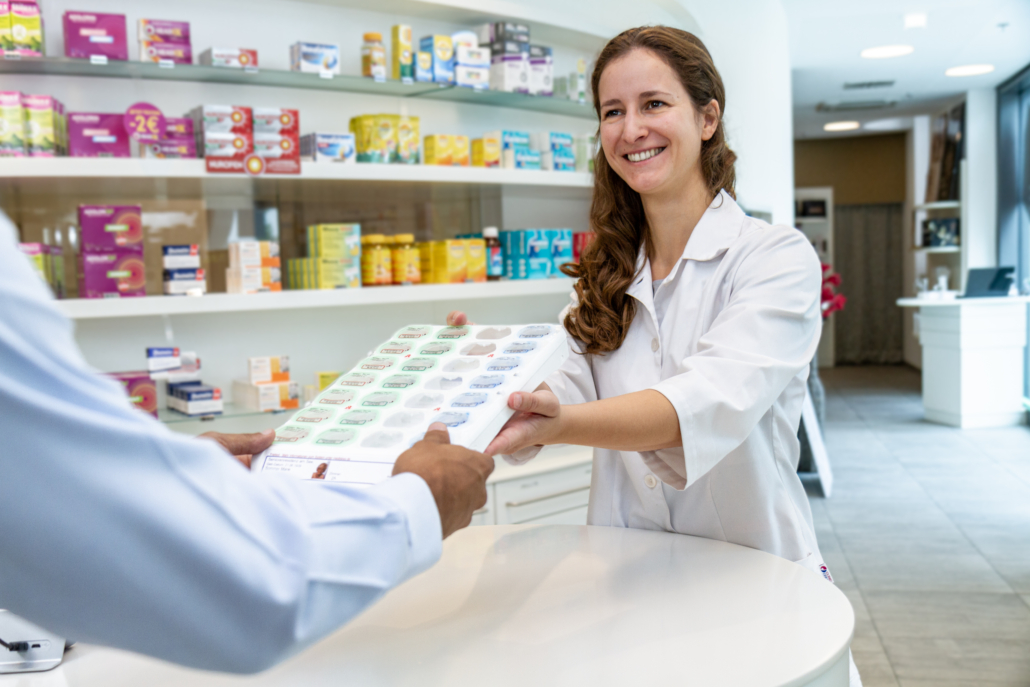 Apothekerin übergibt einem Kunden ein Medikationstablett mit patientenindividueller Verblisterung in einer modernen Apotheke – im Hintergrund Regale mit Medikamenten und Gesundheitsprodukten.