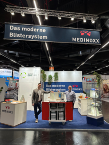 Zwei Mitarbeiter von Medinoxx stehen an einem Messestand auf der ALTENPFLEGE Messe. Hinter ihnen ist ein Banner mit der Aufschrift ‚Das moderne Blistersystem‘ zu sehen. Auf dem Stand werden Blistertrays, ein Siegelautomat und das EcoCircle Recyclingkonzept präsentiert.