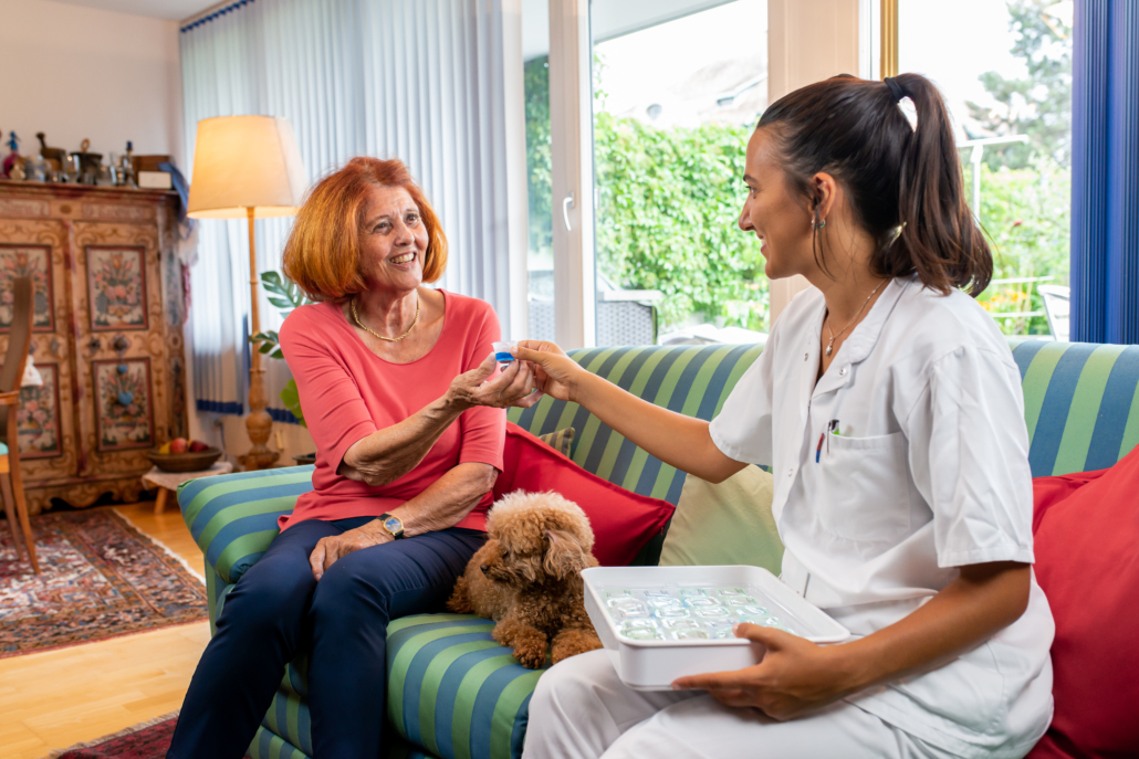 Medinoxx Medikamentenausgabe - Häusliche Pflege Pflegekraft übergibt einer Seniorin einen Medinoxx Medi-Becher in einer häuslichen Umgebung.